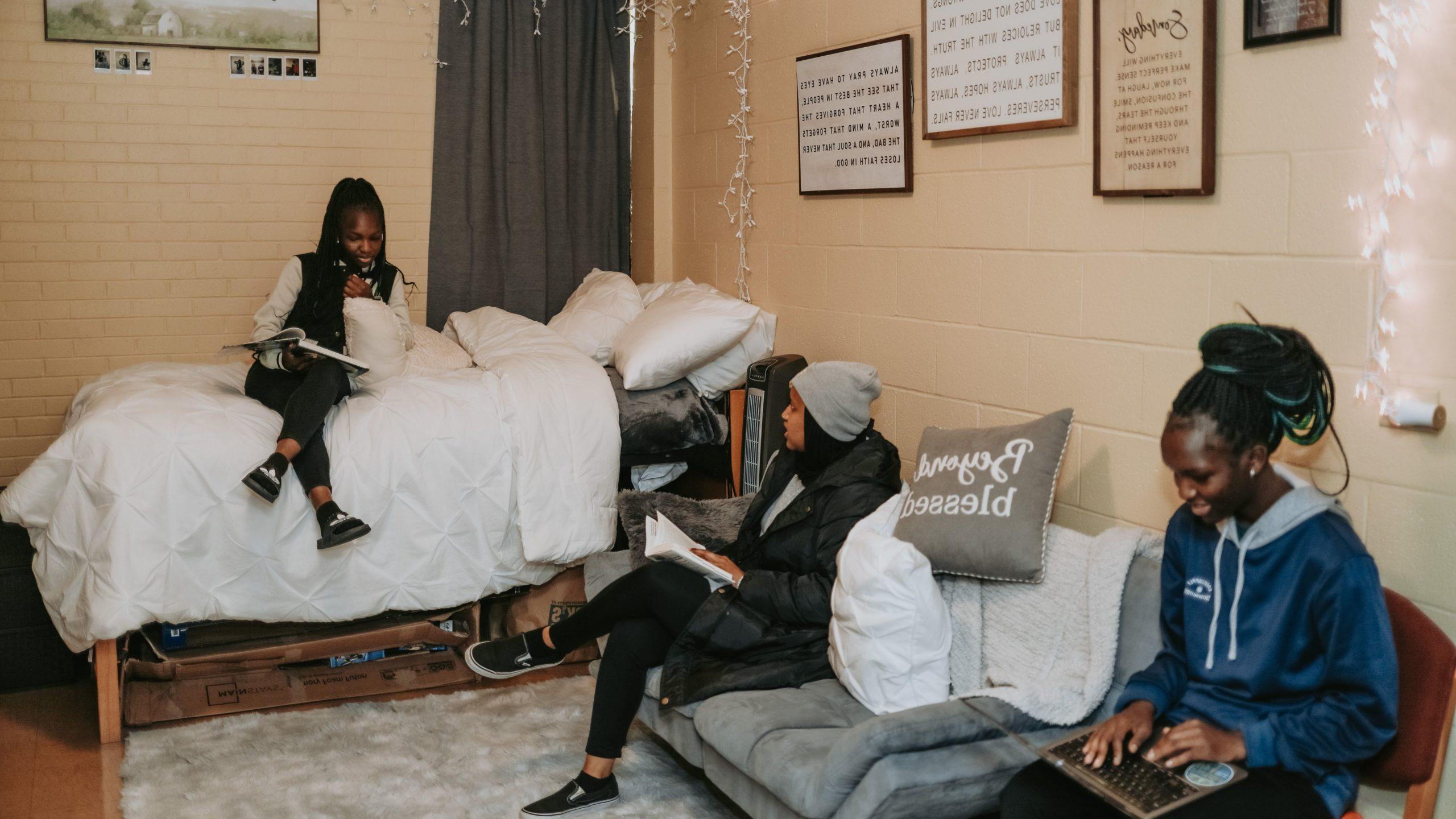 Three students in a dorm room; one is sitting on a bed, 一个在沙发上, 一个人坐在椅子上摆弄着她的笔记本电脑.
